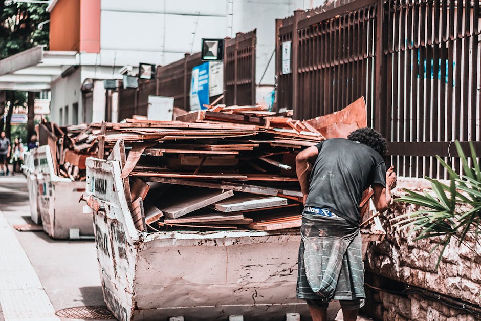 In an urban outdoor setting, a man with dark curly hair, dressed in a black T-shirt and grey checkered shorts, stands beside an overflowing skip filled with construction and renovation waste, includin