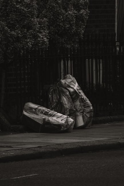 A narrow urban alleyway cluttered with large piles of mixed waste materials, including cardboard boxes, plastic bags, and debris, stacked against the back wall. In the foreground, a large, weathered b